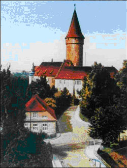 Kolorowy obrazek. Na pierwszym planie droga brukowana prowadząca do zamku. Zamek częściowo zasłonięty przez zielone drzewa.