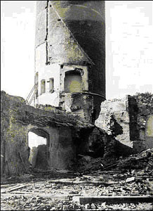 Czarno-białe zdjęcie. Po środku gruzowisko otoczone dwoma sąsiadującymi częściowo rozebranymi ścianami. Za nimi częściowo widoczna okrągła wieża.